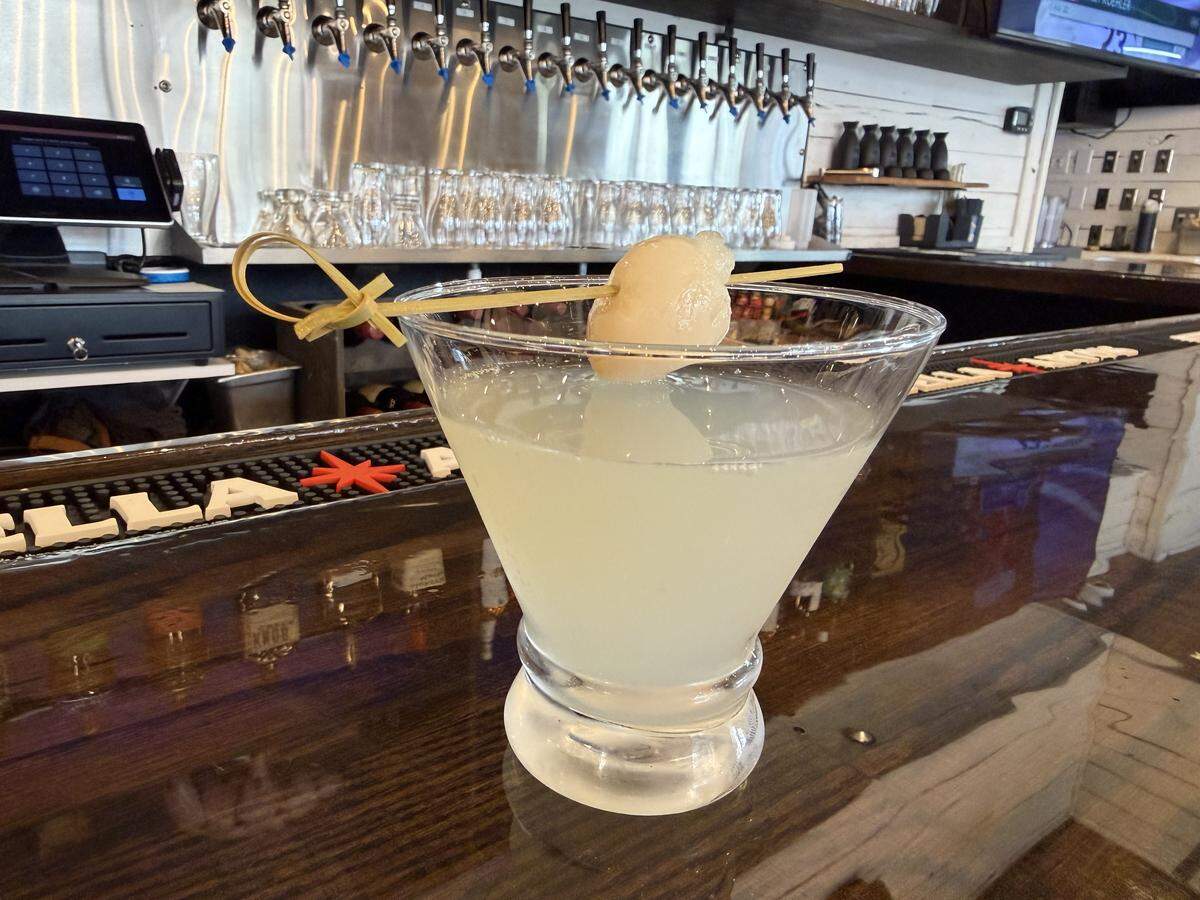 A stemless martini glass filled with a translucent, pale yellow cocktail sits on a polished dark wood bar top. The drink is garnished with a single lychee fruit on a bamboo skewer. In the background, a row of silver beer taps and various glassware are visible in a brightly lit modern bar setting.