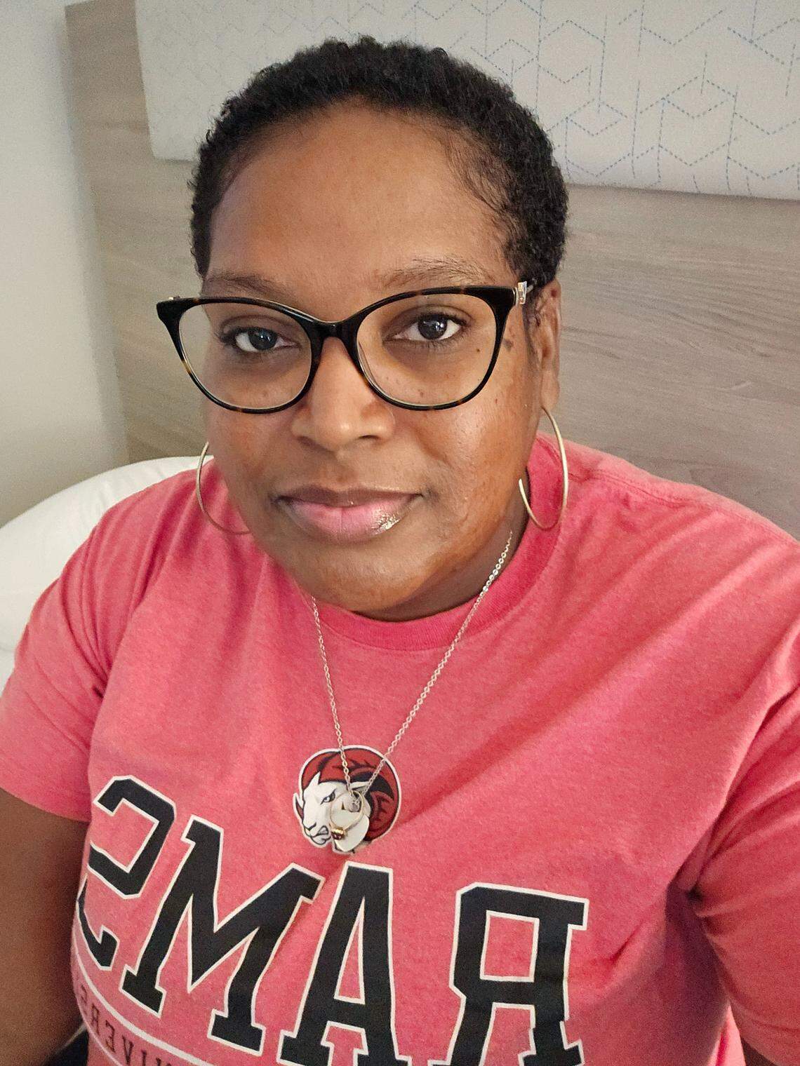 A chest-up portrait of a person with short, dark, curly hair wearing black-rimmed glasses, gold hoop earrings, and a red t-shirt with a white and black logo/text. They are looking directly at the camera.