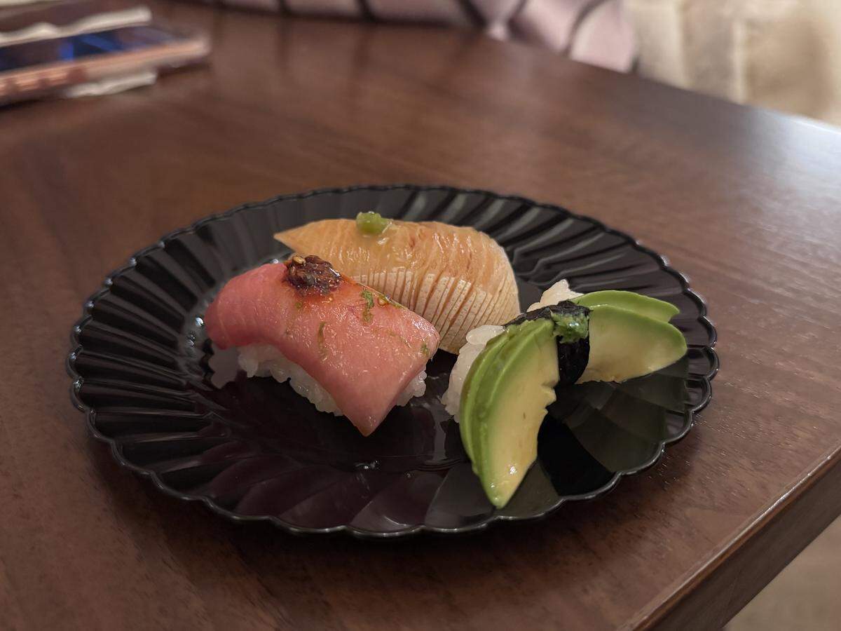 A top-down shot of three pieces of sushi—one tuna with a garnish, one white fish, and one avocado—served on a small, black scalloped plate resting on a dark wooden table.