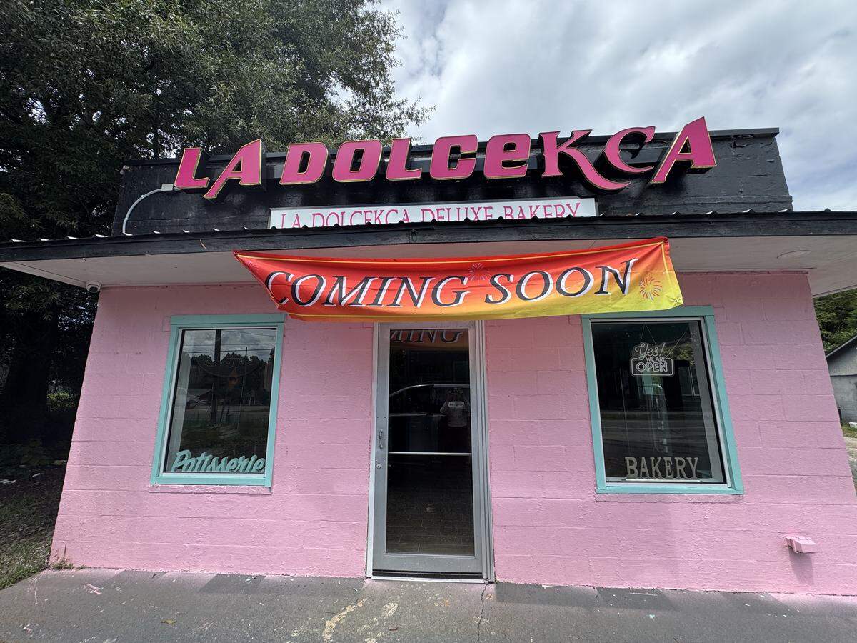 A bright pink building with a black roof and a large red sign that says “LA DOLCEKCA” is shown. Below the main sign, a smaller white sign reads “LA DOLCEKCA DELUXE BAKERY.” A large orange and red banner with the words “COMING SOON” hangs below the signs. The building has two large windows with turquoise frames. The left window has the word “Patisserie” written in white script, and the right window has the word “BAKERY” in white capital letters.
