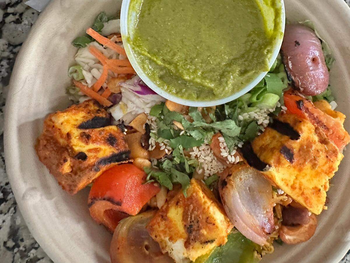 A top-down close-up shows a Botiwalla Desi salad served in a compostable paper bowl. The bowl is filled with a colorful array of ingredients including what appears to be shredded cabbage and carrots as a base. Prominently featured are several pieces of grilled paneer cheese, some with char marks, alongside grilled red and green bell peppers and onions. The salad is garnished with fresh cilantro, sesame seeds, and what look like crushed nuts. At the top of the bowl, a small red potato is visible. A clear plastic cup filled with a vibrant green chutney or dressing is placed on top of the salad, slightly off-center towards the top.
