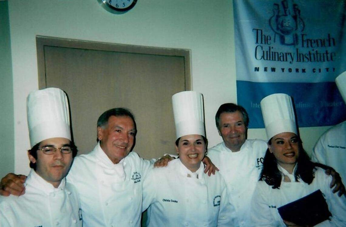 A celebratory group photograph from a culinary school graduation. Five chefs in crisp white uniforms and tall hats pose with their arms around each other. In the center are renowned chefs Jacques Torres and Jacques Pépin, celebrating with three smiling graduates. A blue banner for “The French Culinary Institute New York City” is visible in the background.