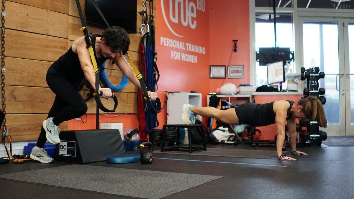 A wide, eye-level shot of two women in a “true 180” gym. On the left, a woman in black workout clothes uses a yellow suspension trainer, straining in a low-lunge position. On the right, another woman holds a high plank on the mat. The background features wood-paneled and bright orange walls with the gym’s “PERSONAL TRAINING FOR WOMEN” logo.