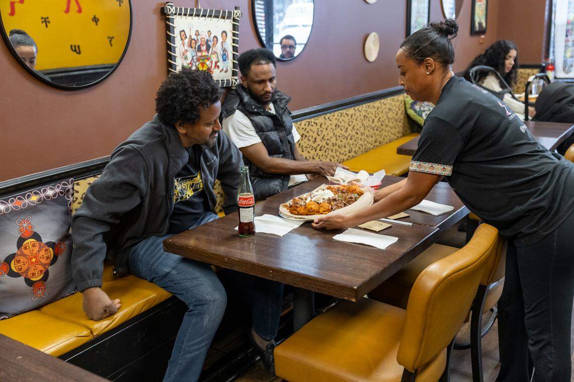 A server places a large, steaming platter of Ethiopian delicacies onto a table for two people. One, wearing a grey fleece, looks on with anticipation. On the table sits a classic glass bottle of Coca-Cola and white napkins. The background wall is decorated with traditional Ethiopian artwork and circular mirrors, contributing to the restaurant’s authentic cultural aesthetic.