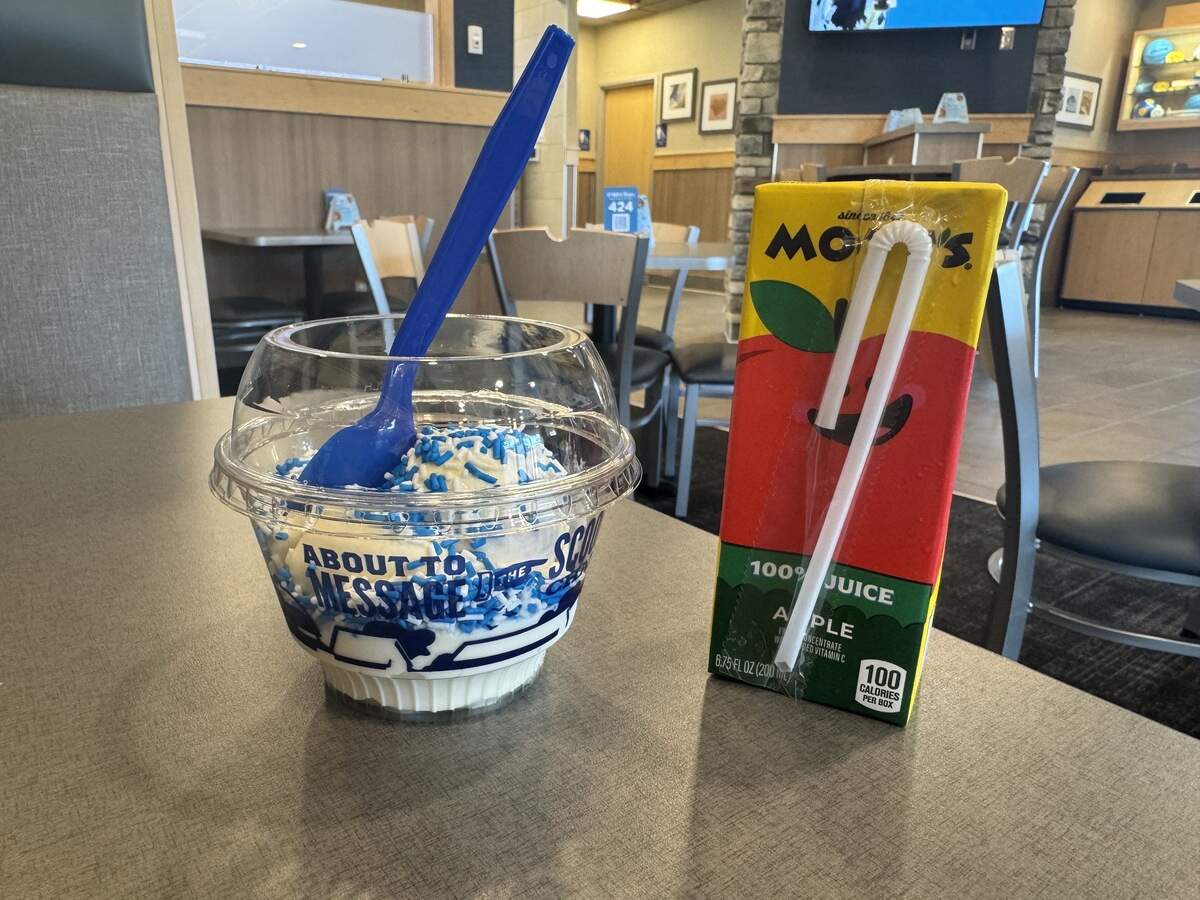 A blue spoon rests inside a clear plastic cup filled with vanilla ice cream and blue sprinkles, sitting on a restaurant table next to a Motts 100% Apple Juice box with a white straw attached. The blurred background shows the interior booths and tables of a Culver’s restaurant.