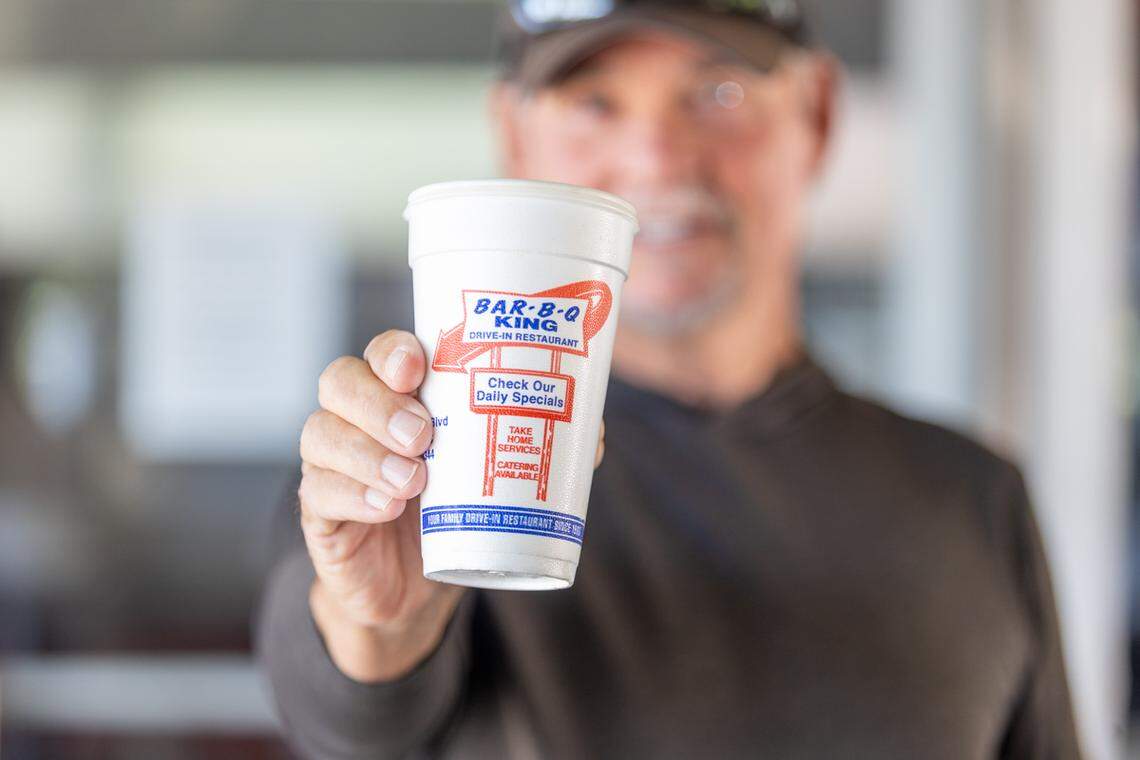 A close-up shot focusing on a hand holding a white styrofoam cup from “Bar-B-Q King Drive-In Restaurant.” The cup’s red and blue logo is in sharp focus, while the person holding it is blurred in the background.
