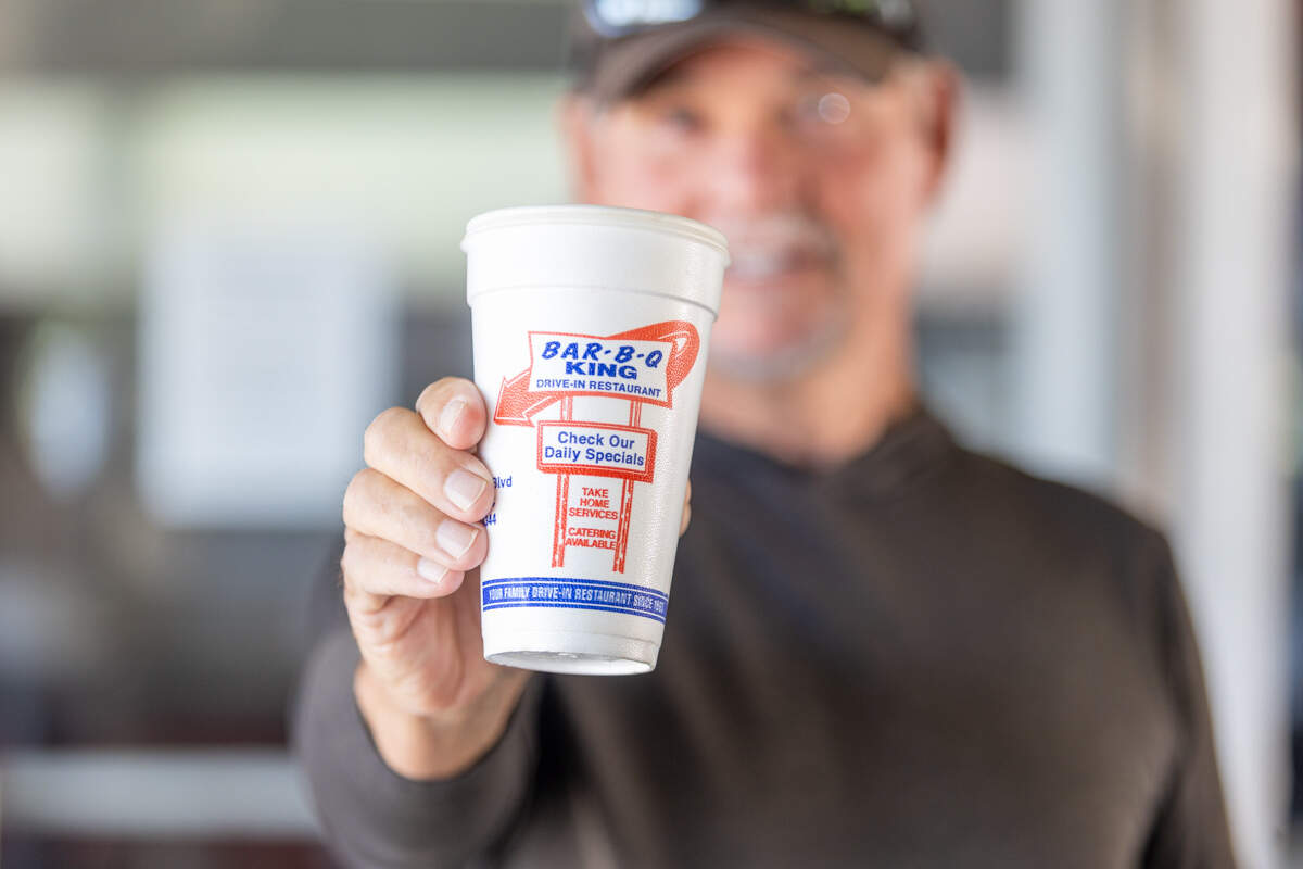 A close-up shot focusing on a hand holding a white styrofoam cup from “Bar-B-Q King Drive-In Restaurant.” The cup’s red and blue logo is in sharp focus, while the person holding it is blurred in the background.