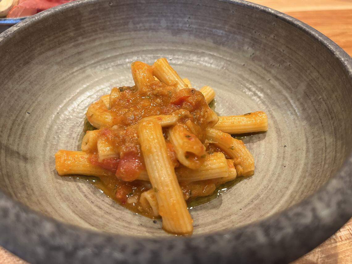 A close-up, high-angle shot of a pasta dish served in a round, rustic gray bowl. The dish features short, tubular pasta (likely rigatoni or ziti) covered in a chunky red tomato sauce with visible herbs and a drizzle of olive oil. The pasta is arranged in a small pile in the center of the bowl, which has a wide, textured rim. The bowl rests on a wooden surface.