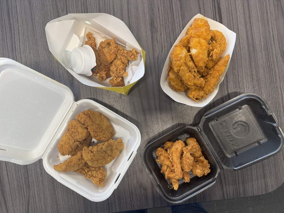 A top-down, overhead view of four different styles of fried chicken tenders arranged in their takeout containers on a dark wood-paneled table. The selection includes chicken in an open yellow-and-white paper box, a small white paper boat, an open white styrofoam container, and a small, open black styrofoam container. Each container holds a varying number of crispy, golden-brown chicken strips.