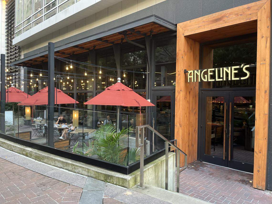 A street-level view of the exterior of a restaurant called “Angeline’s.” The front of the restaurant is a modern design with large wooden panels framing the entrance. The name “ANGELINE’S” is displayed in large, illuminated letters. To the left, there is an outdoor patio with tables, chairs, and several red umbrellas. String lights are hung over the patio, creating a warm ambiance. The patio is surrounded by a glass and metal barrier.