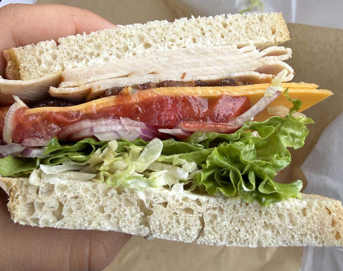 A close-up, cross-section view of a turkey sandwich on white bread. The colorful layers include sliced turkey, yellow cheese, a red tomato relish, sliced red onions, and crisp lettuce. The sandwich is held by a hand over brown wrapping paper.