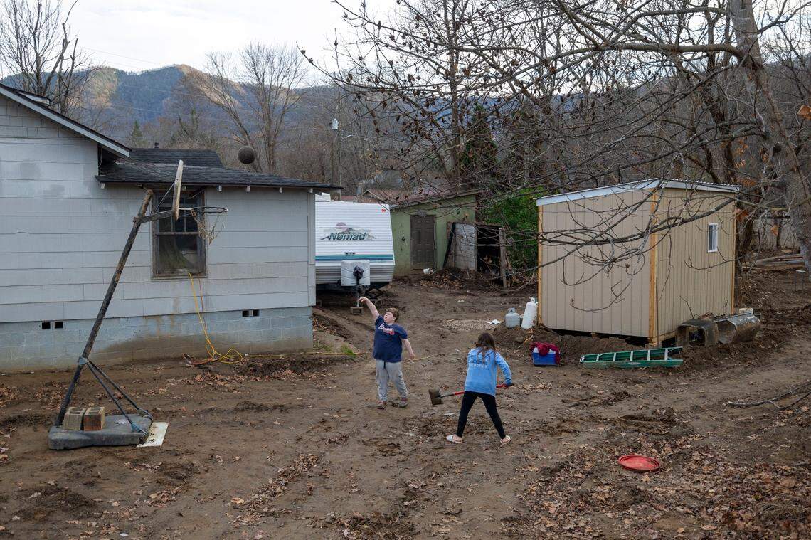 Liam Whiteside, shoots baskets on Wednesday, November 27, 2024, in muddy yard at his grandfather Thomas Whiteside’s home in Black Mountain, N.C. The elder Whiteside has moved into the temporary structure, right, that was built by Cabins 4 Christ on his property after his home was flooded by Hurricane Helene.
