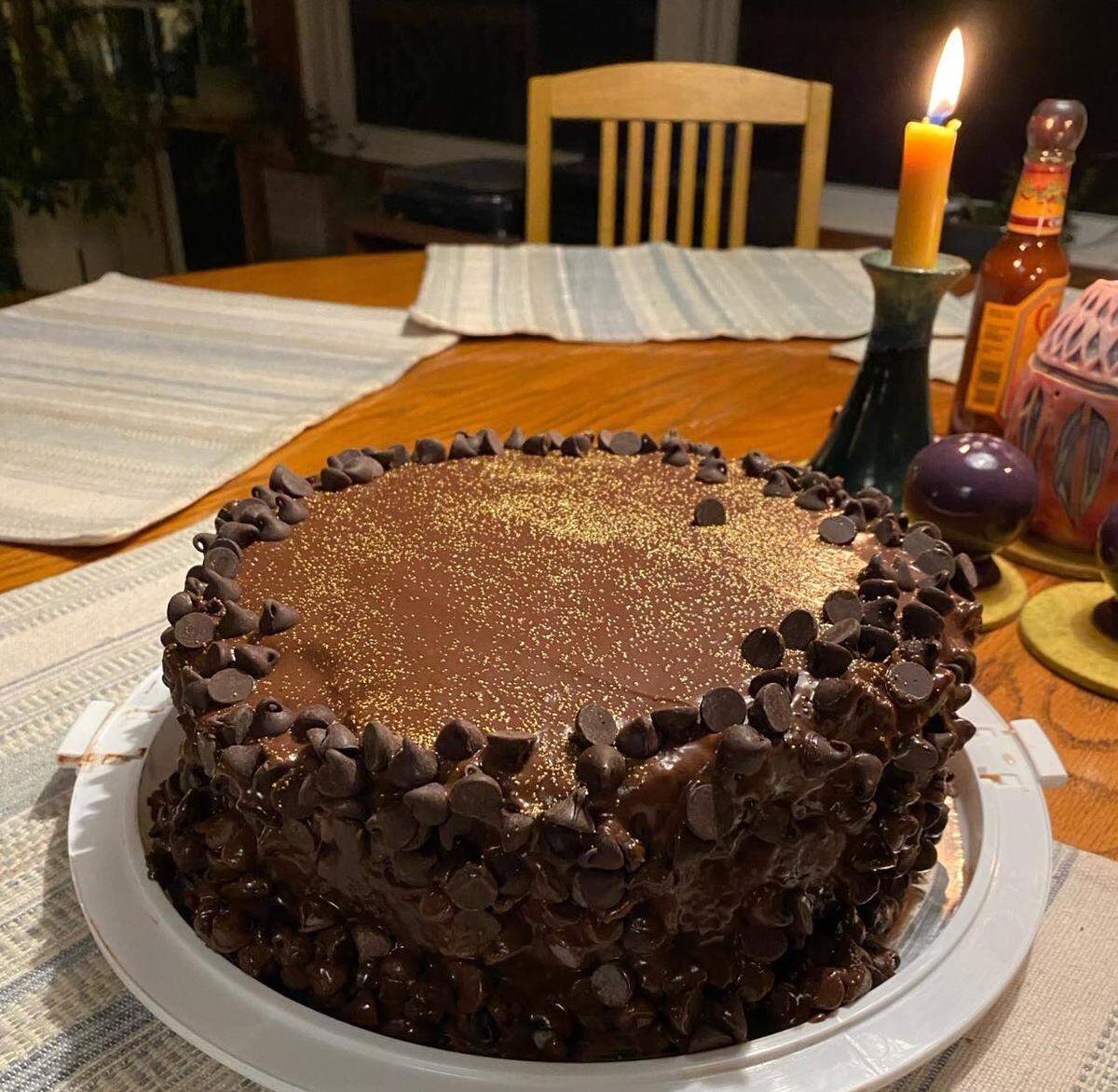 A celebratory scene featuring a homemade chocolate cake on a white platter. The decadent cake is coated in a glossy chocolate ganache, with its sides and top border encrusted with chocolate chips and the top surface dusted with edible gold glitter. The cake is on a dining room table, illuminated by the warm flame of a single candle.