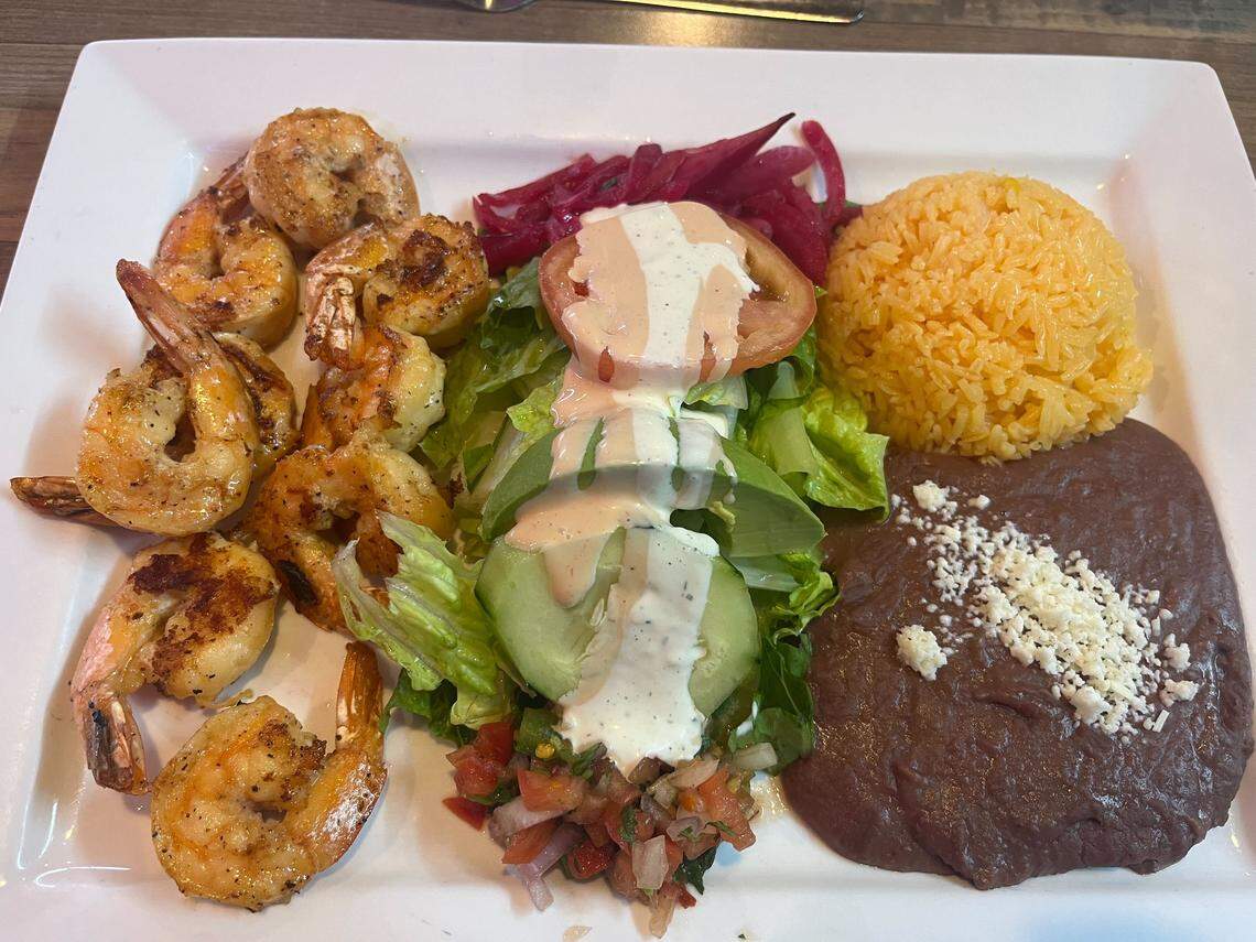 A plate of food featuring several grilled, seasoned shrimp on the left, next to a bed of lettuce topped with sliced avocado, pico de gallo, and a slice of tomato drizzled with white dressing. The plate also includes a scoop of yellow rice and a portion of refried beans topped with crumbled white cheese, with a side of pickled red onions.