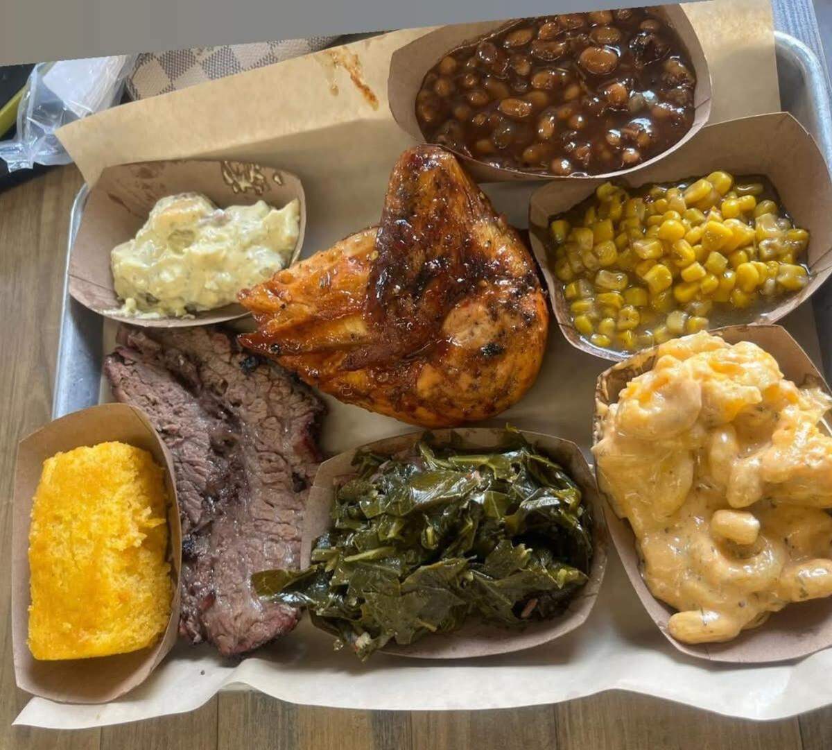 A large metal tray lined with butcher paper featuring a variety of BBQ dishes served in individual cardboard boats. The platter includes sliced brisket, a roasted chicken quarter, collard greens, mac and cheese, baked beans, corn, potato salad, and a square of cornbread.