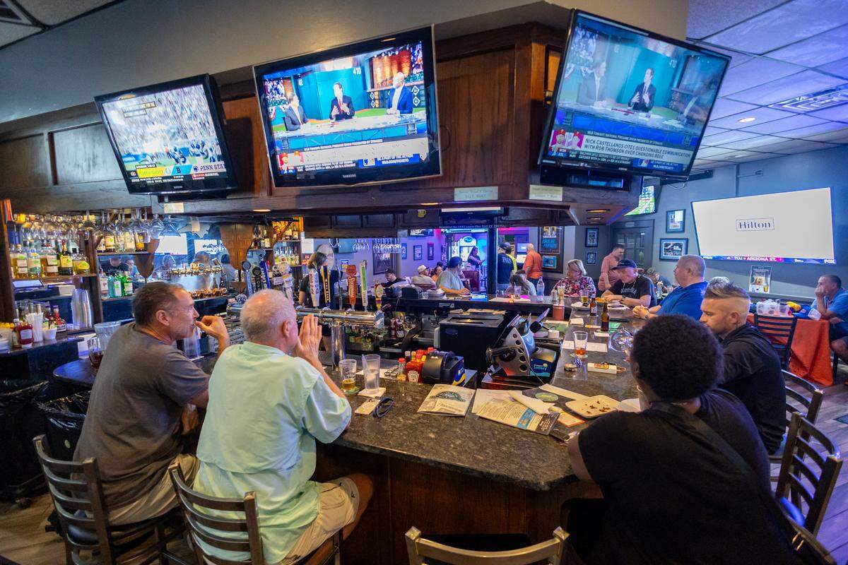 A candid shot of the lively atmosphere at the Sports Page bar. A diverse group of patrons sits along the dark granite bar, engaged in conversation and watching the several large TVs showing sports. The bar is well-stocked, and the lighting is typical of a casual sports bar.