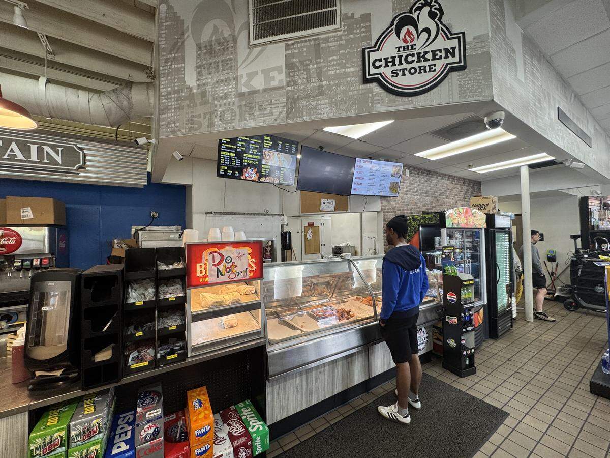 An interior view of a casual fast-food restaurant counter named “The Chicken Store.” A customer in a blue hoodie and black shorts stands looking into a long glass display case filled with hot food. Above the counter are digital menu boards and a large circular logo featuring a black rooster silhouette. Boxes of soda cans, including Mountain Dew, Pepsi, and Sprite, are stacked on the floor beneath the counter area.