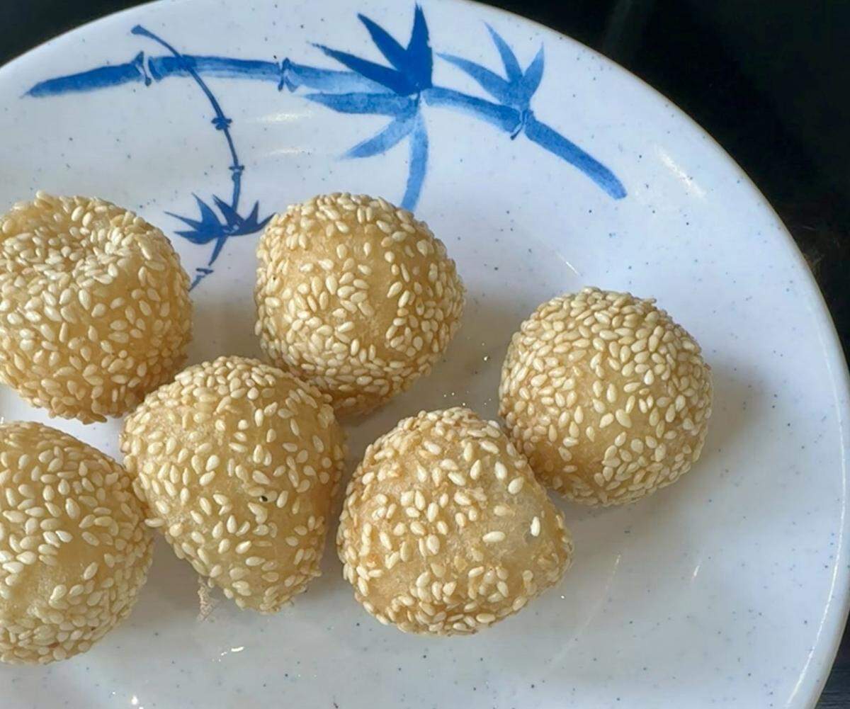 A plate of six Chinese sesame balls from LanZhou. The deep-fried pastries have a golden hue, are generously coated in white sesame seeds, and are filled with red bean paste, as per the file name. They are presented on a white plate with a blue decorative motif.