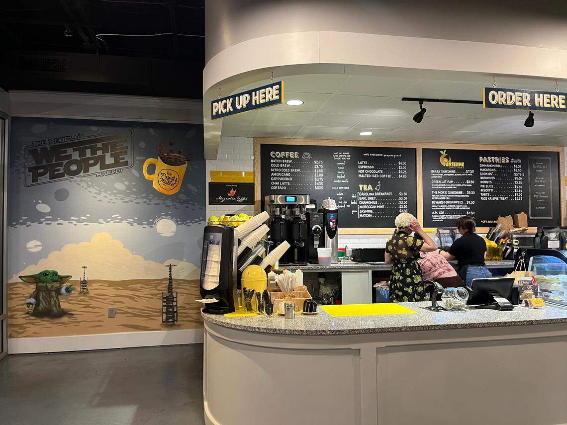 An eye-level, medium shot of a coffee shop’s counter area. To the right is the curved ordering counter with “ORDER HERE” and “PICK UP HERE” signs above, and chalkboard menus on the back wall. To the left is a large wall mural featuring a Star Wars theme with Grogu (Baby Yoda) on a desert planet and the text “WE THE PEOPLE... fuel.”