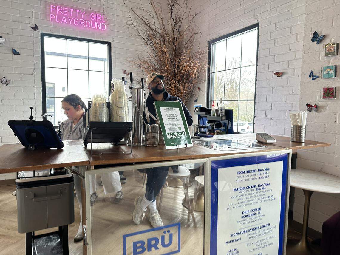 A wide shot of a modern, minimalist coffee bar named “BRÜ” inside a bright room with white brick walls. A barista in a baseball cap and hoodie stands behind a wooden counter, looking upward, while a staff member works to the left. A pink neon sign reading “PRETTY GIRL PLAYGROUND” glows on the wall. The menu on the front of the counter lists “Draft Oat Latte” and “Matcha on Tap.”