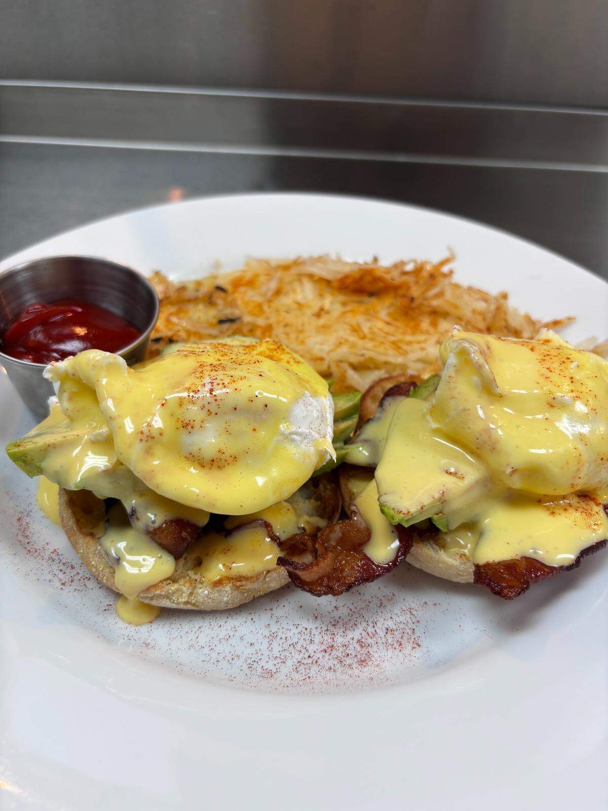 A plate of Eggs Benedict with avocado and bacon on an English muffin, covered in hollandaise sauce and served with a side of crispy hash browns and ketchup.