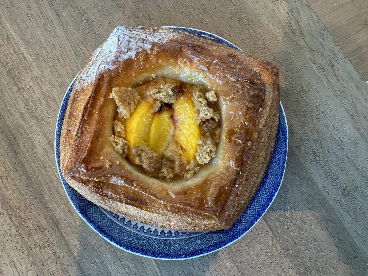 A close-up, top-down shot of a square fruit danish pastry served on a blue and white geometric patterned plate, sitting on a wooden surface. The pastry features a golden-brown, glossy, flaky crust with a light dusting of powdered sugar on one corner. The center is filled with bright yellow baked peach or apricot slices, topped with a light sprinkle of crumbly streusel topping.