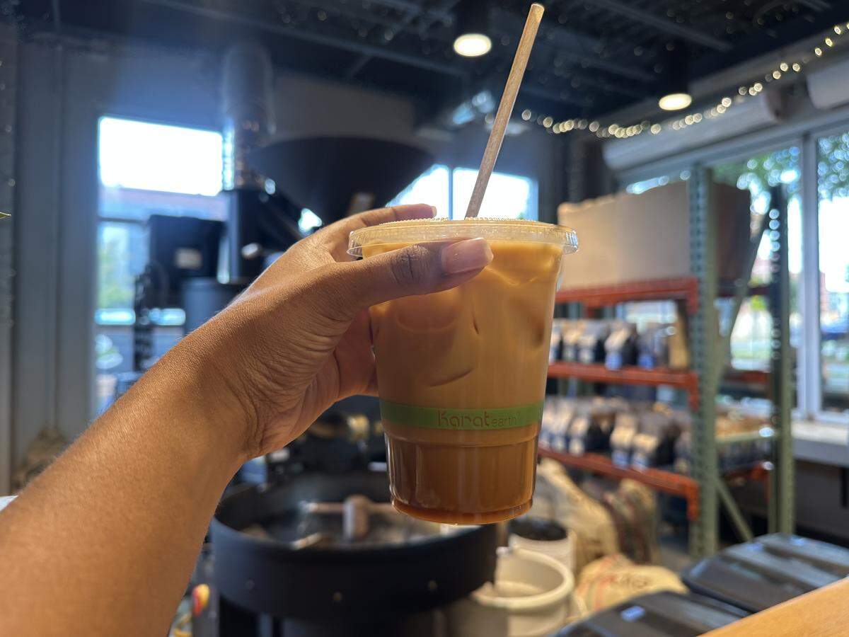A close-up, first-person shot of a hand holding up a clear plastic cup of iced coffee. The drink is a creamy tan color with a darker layer of syrup or coffee at the bottom, and a paper straw sticks out of the lid. The background is blurred, showing the interior of a coffee roastery, with a large coffee roasting machine visible behind the cup and shelves of coffee bags to the right.