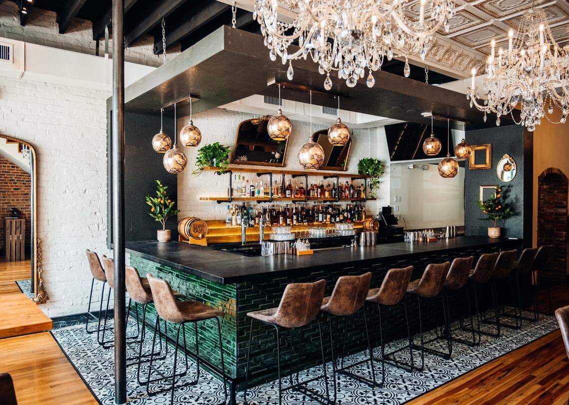 This is an interior shot of a stylish, upscale restaurant bar with a dark, L-shaped countertop and a front clad in glossy green subway tiles. Several ornate crystal chandeliers hang from a tin ceiling, illuminating the bar and the rows of brown leather barstools.