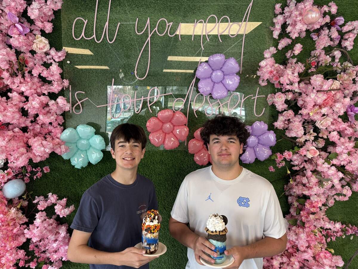 Two teenagers hold gourmet milkshakes in front of a green turf wall decorated with pink blossoms and balloon flowers.
