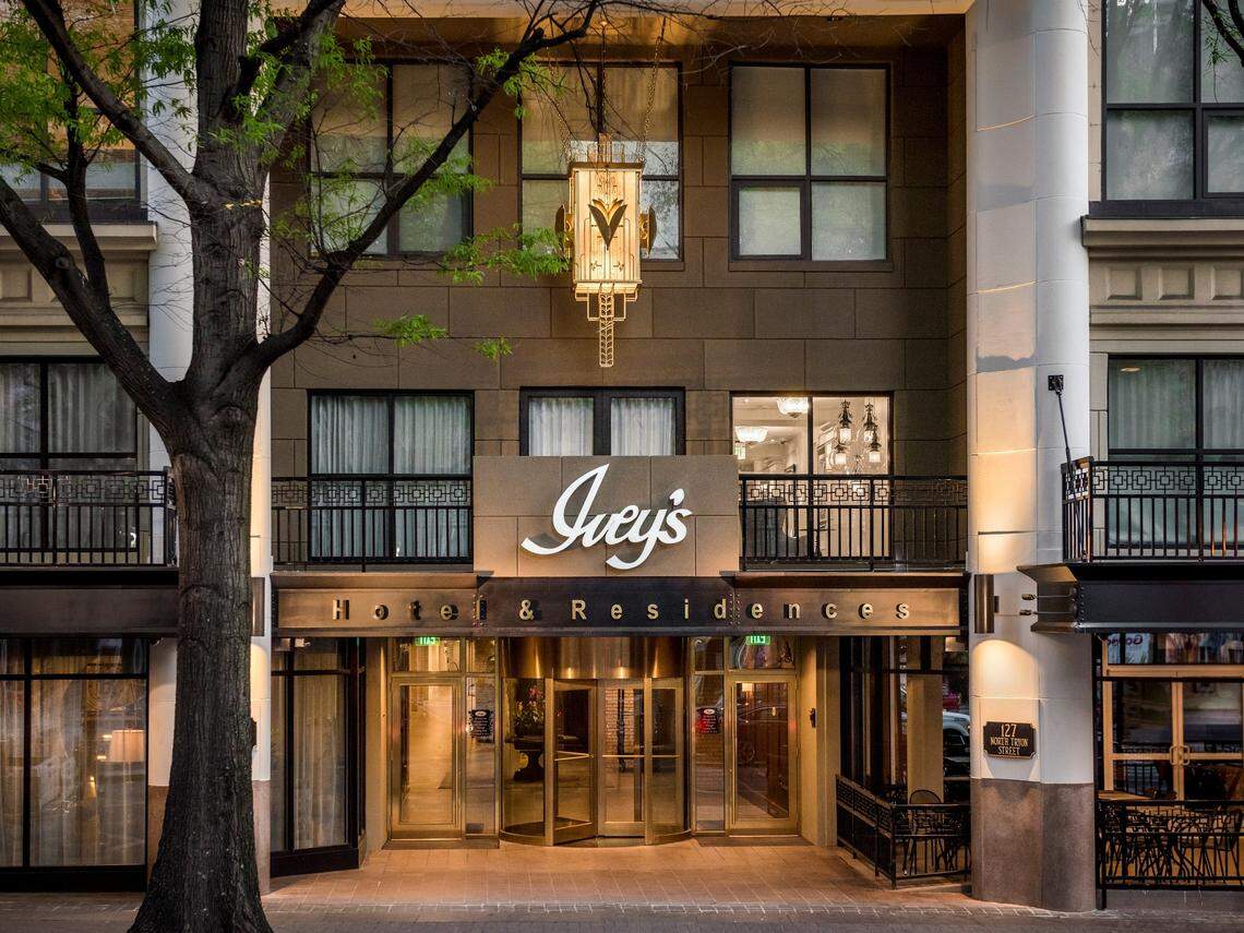 An eye-level exterior photo of the Ivey’s Hotel & Residences entrance. The building facade is made of brown stone, with a large, ornamental, art-deco style lamp hanging over the revolving glass doors. The Ivey’s logo is prominently featured in a white script above the entrance, with a black awning underneath stating “Hotel & Residences.” The entrance is flanked by large white columns and a tree with green leaves on the left.