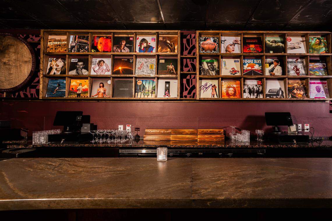 A direct, eye-level view behind the bar, focusing on the extensive vinyl record display. Two large wooden grids are filled with various album covers, ranging from rock to soul, serving as the main backdrop for the bartenders. The bar surface in the foreground is a dark, polished stone, featuring a single small candle and organized stacks of glassware and POS systems at either end.