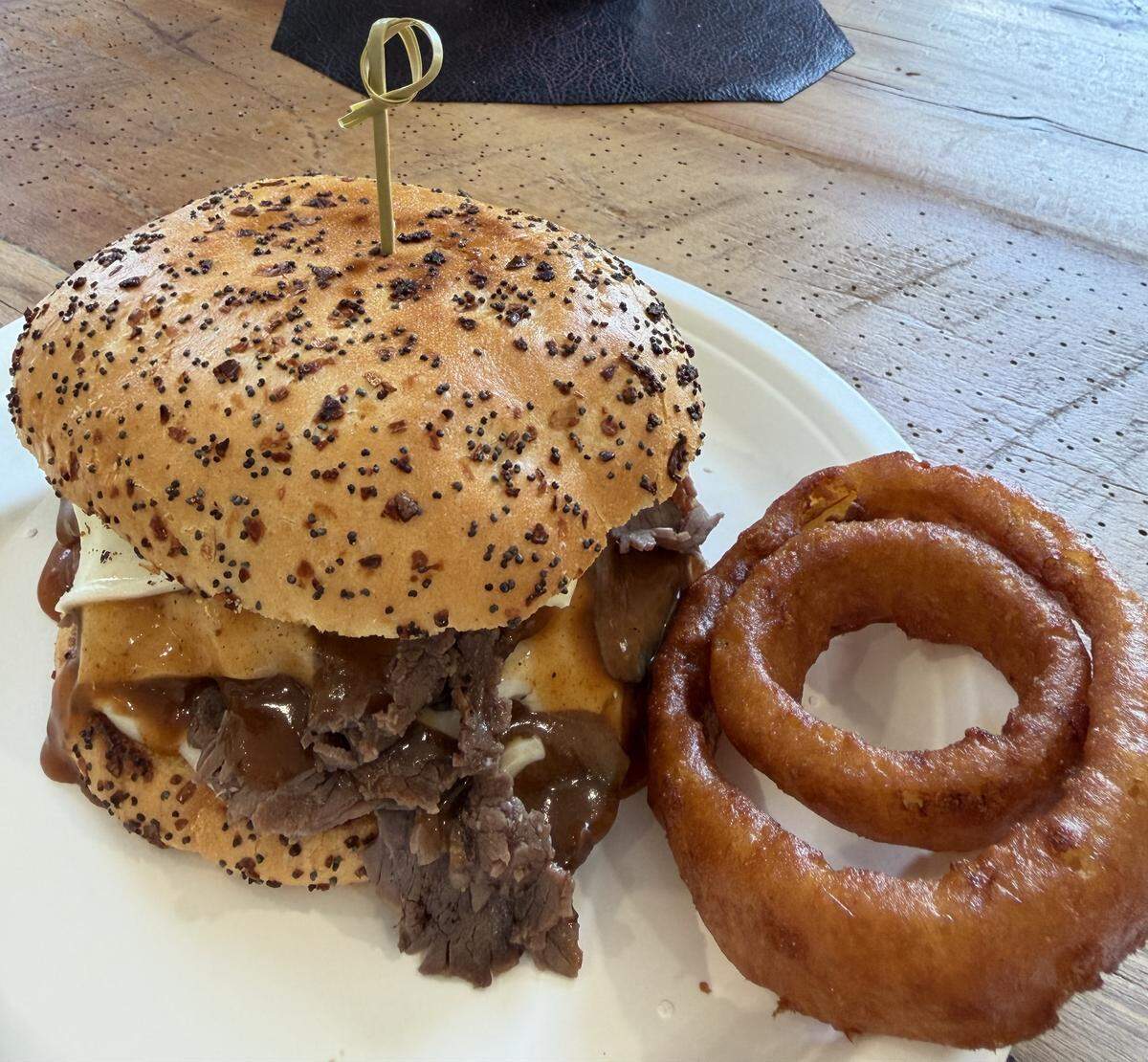 high-angle shot of a large roast beef sandwich on a white plate. The bun is densely covered in poppy seeds and dried onion bits. Thick brown gravy and slices of roast beef spill out from under the bun. Next to the sandwich is a jumbo, thick-cut onion ring with a deep golden, crispy batter.