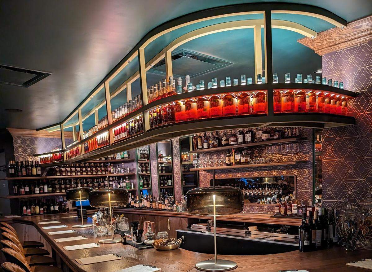 The interior of a chic, moody whiskey lounge. A custom-built, curved shelving unit hangs from the ceiling, its shelves of whiskey bottles brilliantly backlit. The long bar below is set with menus and stylish, mushroom-shaped lamps. The walls are covered in a purple and gold Art Deco-style wallpaper.