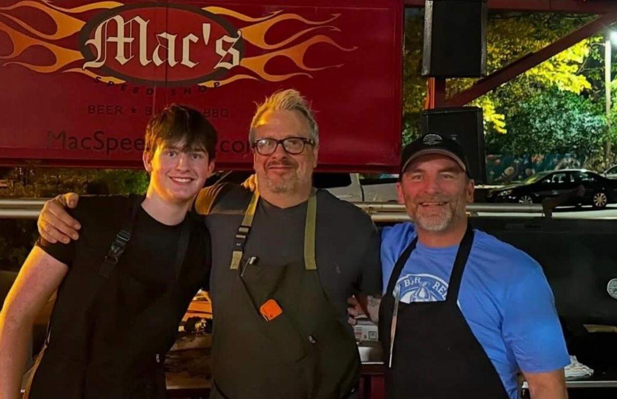 Three chefs standing together at night, posing for a photo with their arms around each other. The one on the left is younger with dark hair, the middle one has grey hair and glasses, and the one on the right is wearing a blue shirt and a baseball cap. They are standing in front of a red sign with “Mac’s Speed Shop” written in stylized text surrounded by orange flames.