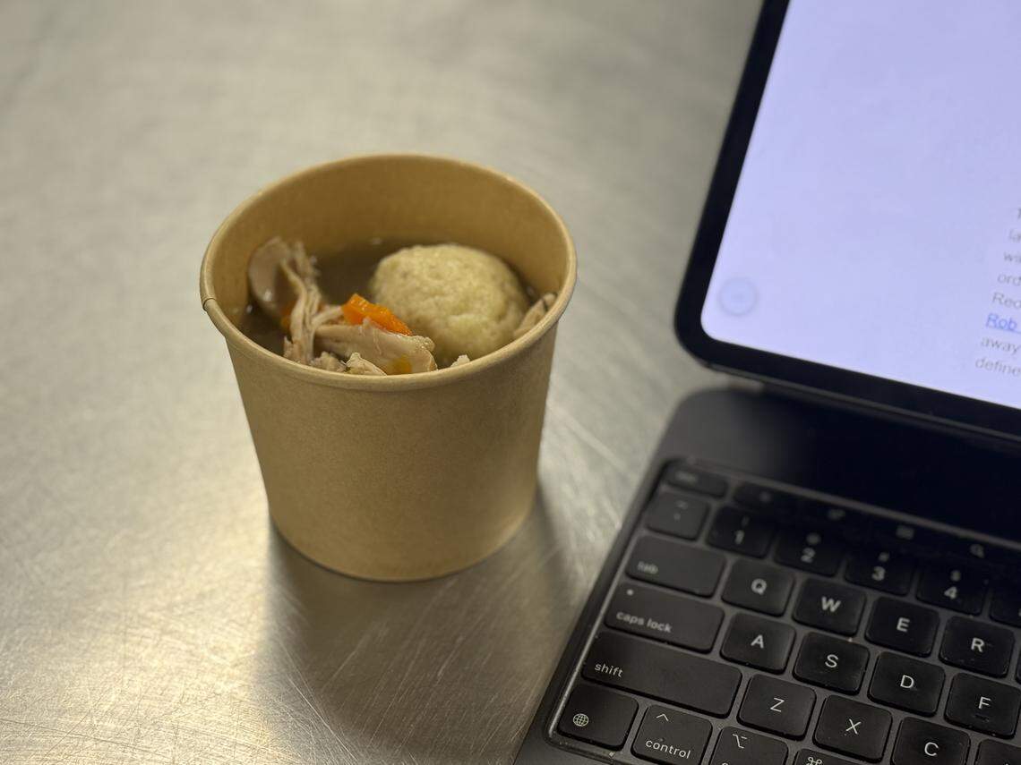 A single serving of matzo ball soup in a brown paper cup sits on a stainless steel table next to a black laptop keyboard and tablet. The soup contains a large, fluffy matzo ball, shredded chicken, and sliced carrots in a clear broth.