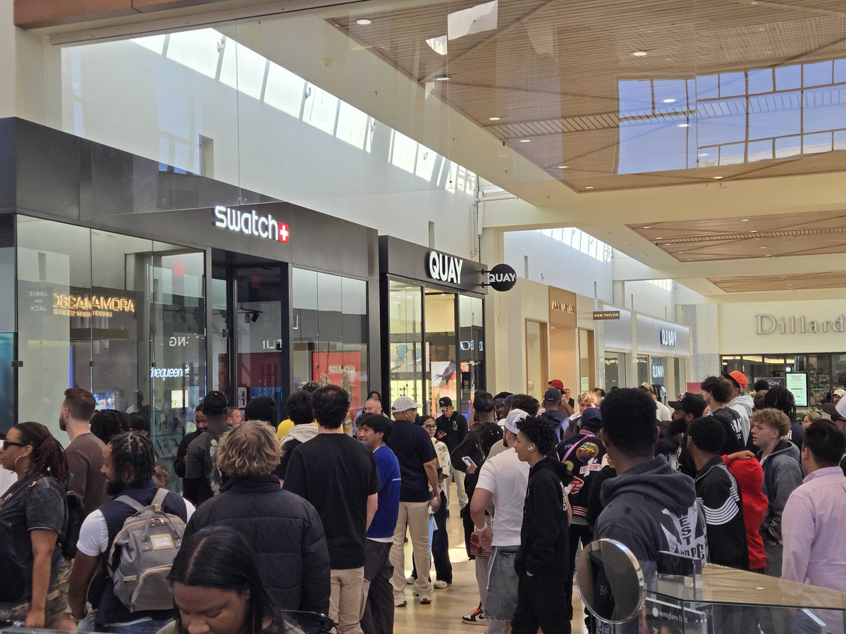 A crowded indoor shopping mall hallway filled with a large group of people waiting in a long line. The crowd consists mostly of young adults dressed in casual clothing like hoodies, t-shirts, and baseball caps. They are gathered in front of a storefront with a black facade and a glowing white and red “Swatch” sign. Next to Swatch is a store named “QUAY” with a black storefront. Further down the brightly lit mall corridor, other store signs are visible, including “Ann Taylor,” “Old Navy,” and a large “Dillard’s” department store entrance in the background. The mall features high ceilings with wooden paneling and large skylights letting in natural light.