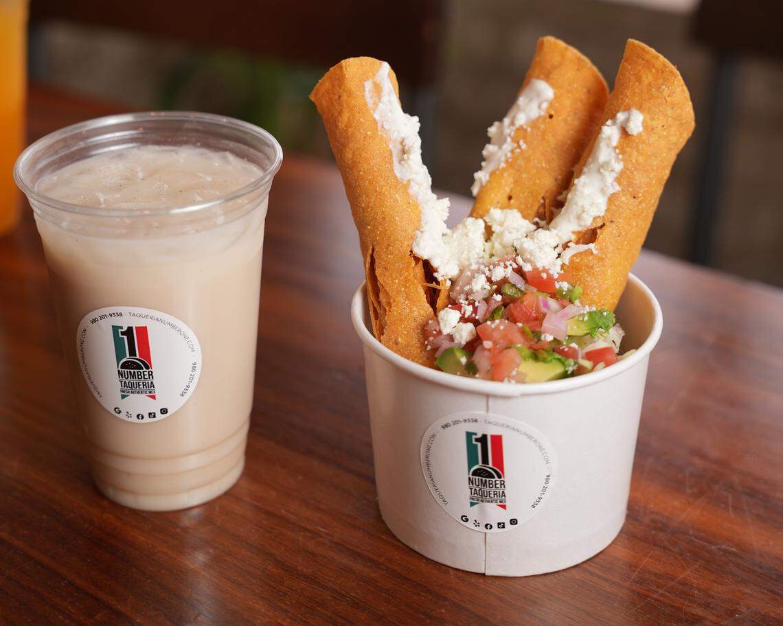 Three taquitos topped with pico de gallo and crumbled white cheese are served upright in a small white paper cup on a dark wooden table. A clear plastic cup of a creamy, light-brown beverage, both featuring a “Number Taqueria” logo sticker, sits next to the taquito cup.