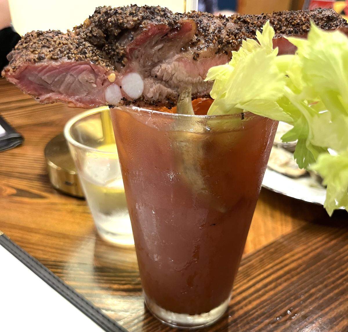 In a dimly lit restaurant, a pint glass is filled with a red Bloody Mary. The cocktail is extravagantly garnished with a whole smoked pork rib and a large celery stalk. The cross-section of the rib shows the pink meat and the thick, peppery bark.