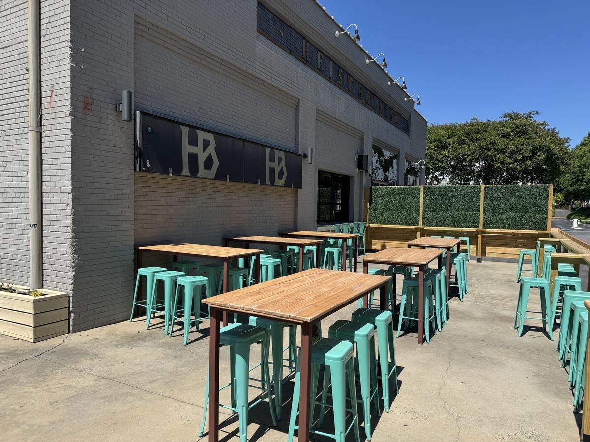 An outdoor patio area at Heist Brewery on a sunny day. Several tall, rectangular wooden tables are paired with bright turquoise metal barstools. The patio is concrete, bordered by a gray brick wall on one side and a wooden fence with a green faux-hedge topper on the other. Black signage with gold “HB” logos is mounted on the brick wall.