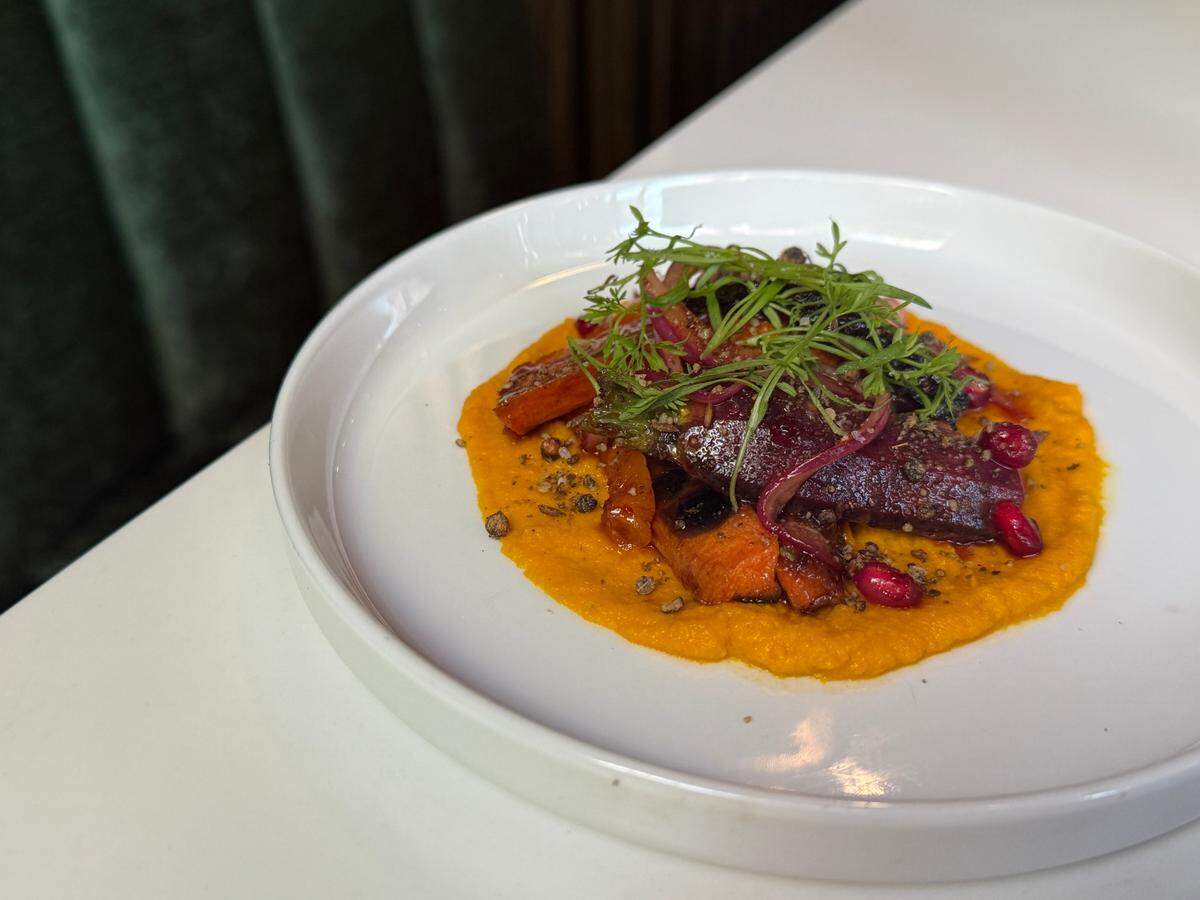 A close-up of a plated dish in a shallow white bowl. A bright orange carrot purée is spread on the plate, topped with roasted carrots, red pomegranate seeds, and a garnish of fresh green micro-herbs.