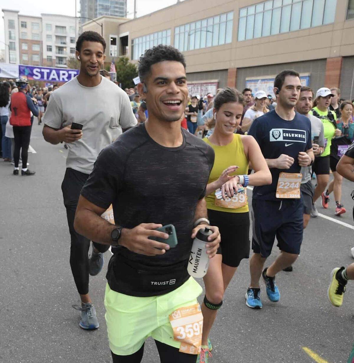 A group of runners participates in a city race, with a smiling runner in a black shirt and neon shorts in the foreground.