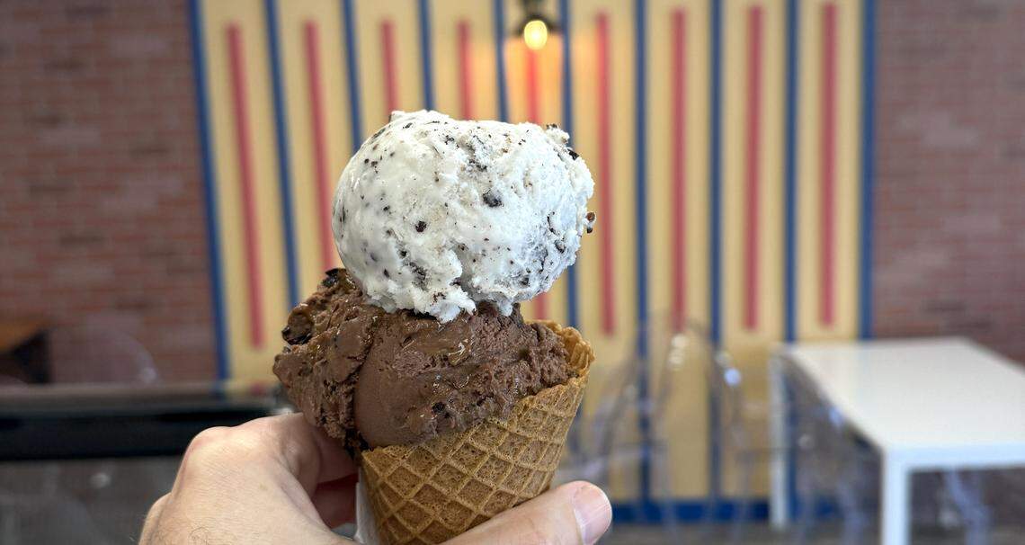 A hand holds a crisp waffle cone topped with two generous scoops of vegelato. The bottom scoop is a rich, dark chocolate, while the top scoop is a creamy white with dark chocolate flecks. The background shows a modern dining area with a brick wall and a colorful striped accent wall.
