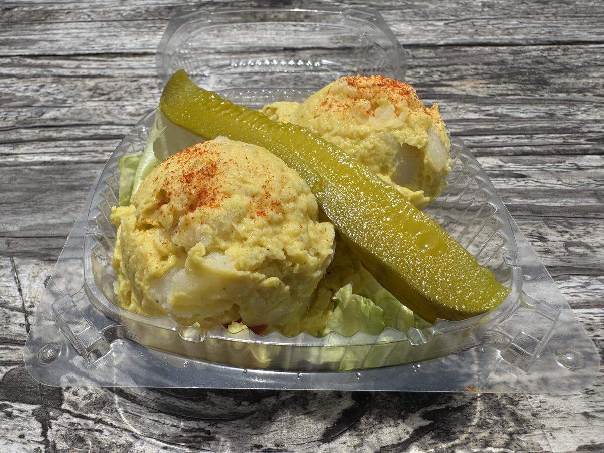 A close-up shot of potato salad served in a clear plastic takeout container on a weathered wooden table. Two large scoops of yellow potato salad are dusted with paprika, nestled on a bed of green lettuce, and topped diagonally with a long, vibrant green dill pickle spear.