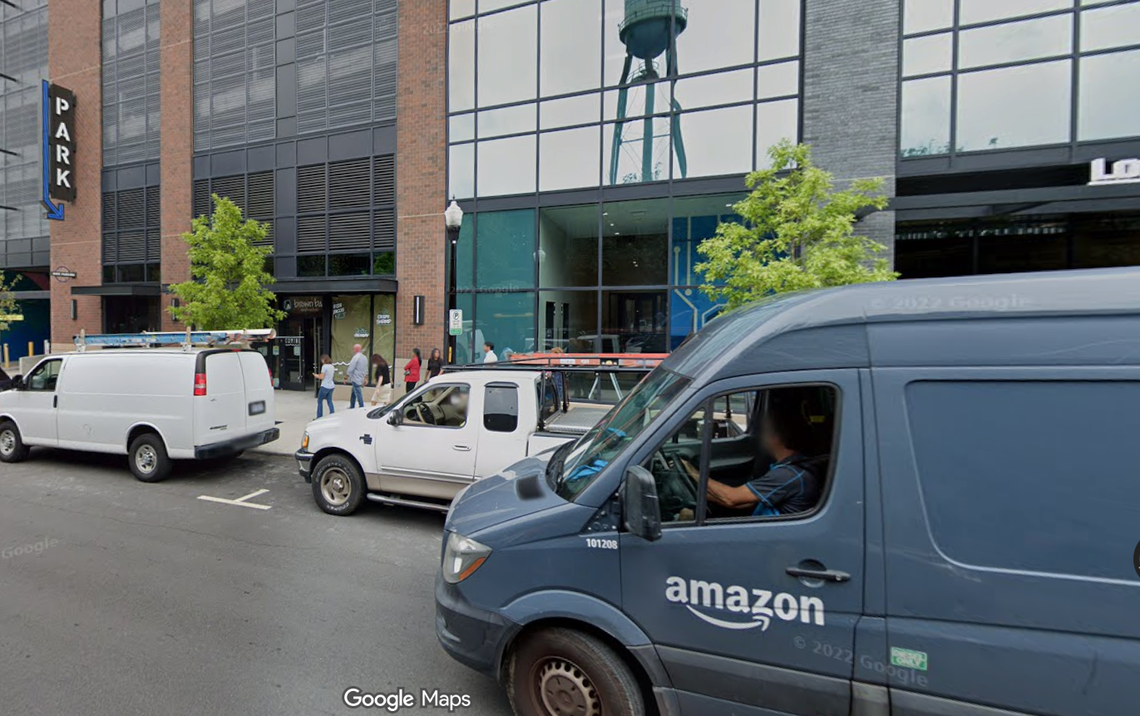 A street view shows a large, dark blue Amazon delivery van parked in the foreground, obscuring the driver’s face, with a white pickup truck and a white cargo van parked behind it along a curb. The background features a modern, multi-story brick building with large glass windows and a water tower visible above the roofline, with a sign that reads “PARK” on the left.