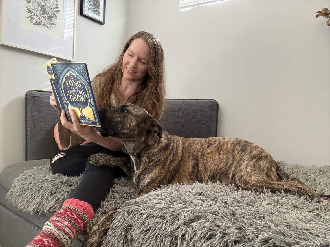 A woman with long reddish hair is sitting on a couch covered with a gray, shaggy blanket, reading the book “As Long as the Lemon Trees Grow.” A brindle dog is lying next to her, looking toward the book.