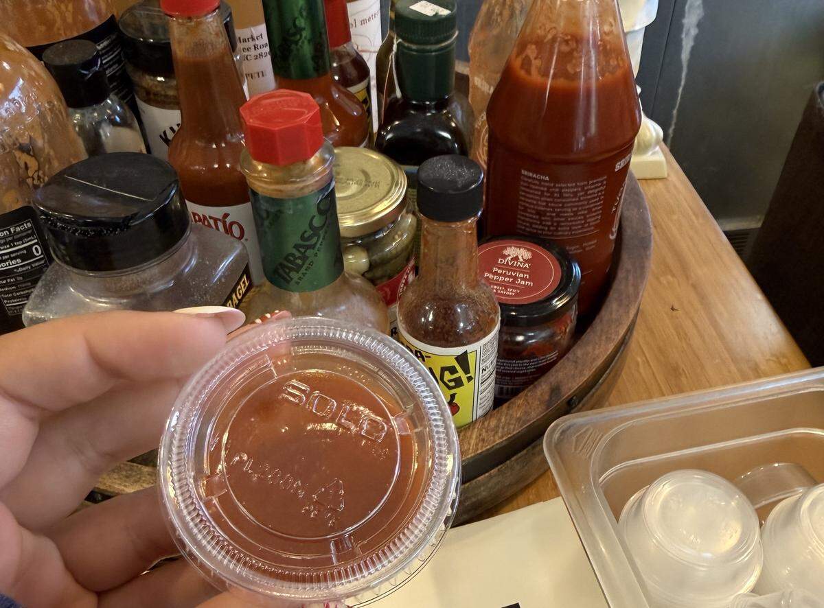 A person’s hand holds up a small, clear plastic portion cup of red sauce with “SOLO” embossed on the lid. In the background, a wooden caddy on a table is filled with an assortment of hot sauce bottles and other condiments.