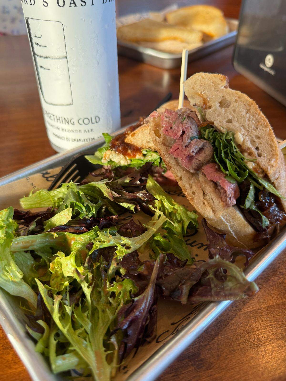 A tray holds a meal consisting of a sandwich cut in half, a side salad, and a can of beer. The sandwich is on crusty bread and appears to be filled with sliced roast beef and greens.