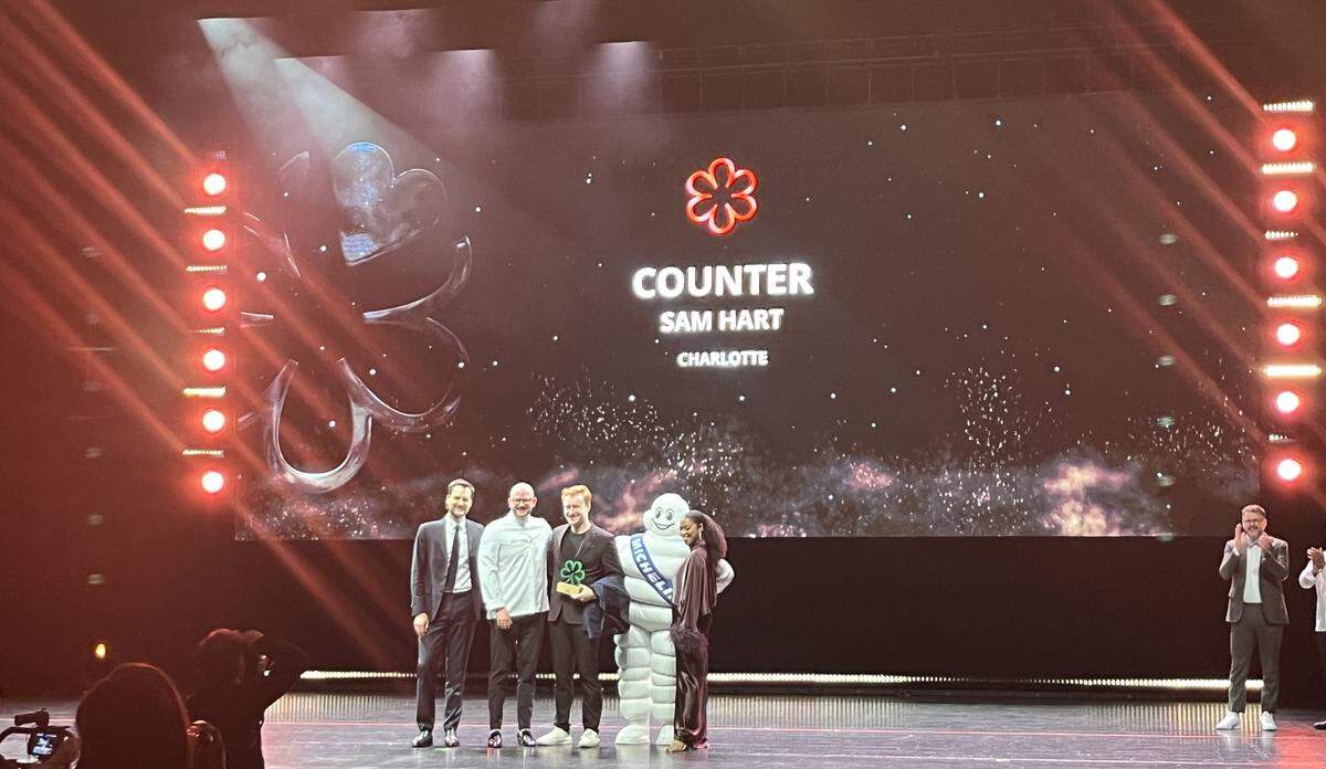 A wide shot from the audience of an awards ceremony on a brightly lit stage. A large screen in the background displays a flower-like logo and the text “COUNTER,” “SAM HART,” “CHARLOTTE.” In the center of the stage, chef Sam Hart smiles as they hold a green award trophy, standing between a host with a microphone and the Michelin Man mascot.