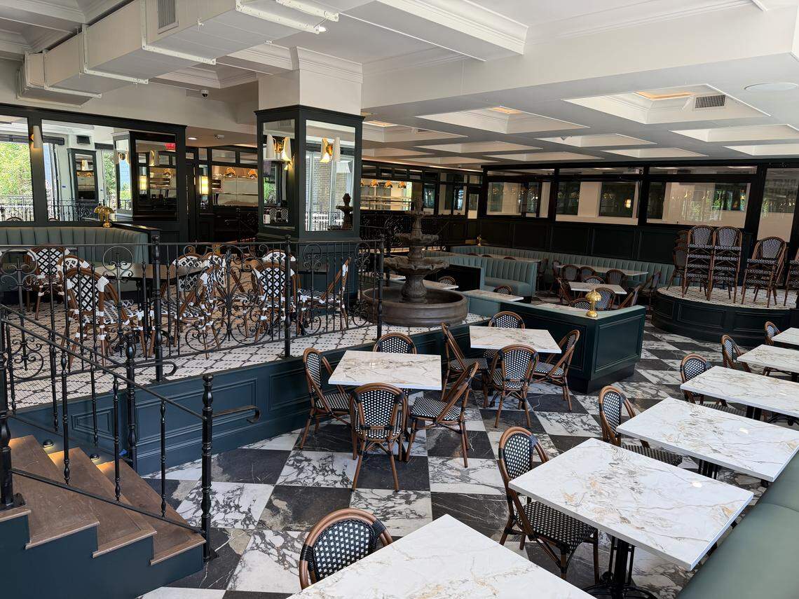 A high-angle view of a split-level dining room featuring black-and-white checkered marble floors on the lower level and intricate tile on the upper level. The space is decorated with teal accents, marble-topped tables, and a central tiered stone fountain.