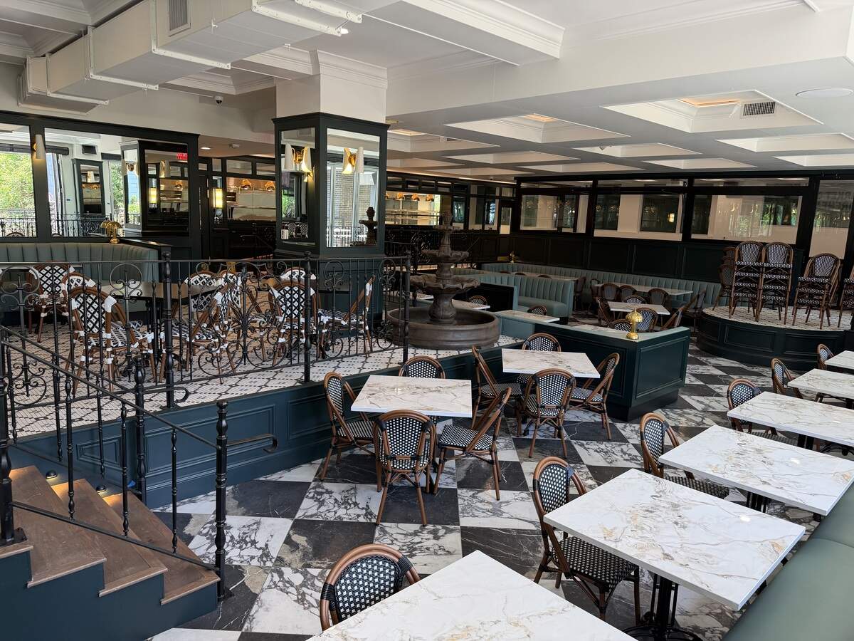 A high-angle view of a split-level dining room featuring black-and-white checkered marble floors on the lower level and intricate tile on the upper level. The space is decorated with teal accents, marble-topped tables, and a central tiered stone fountain.